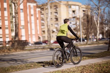 Orta yaşlı bir kadının arka görüntüsü, bir şehir caddesinde bisiklet sürüyor..
