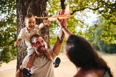 Happy family playing in nature with a toy airplane while father holds his son on his shoulders.  clipart