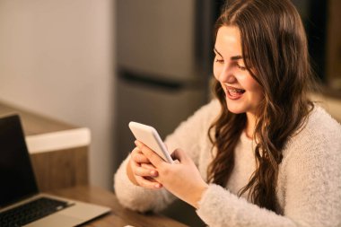 Telli neşeli genç bir kadın, evinde oturmuş, akıllı telefonuyla video sohbeti yapıyor.. 