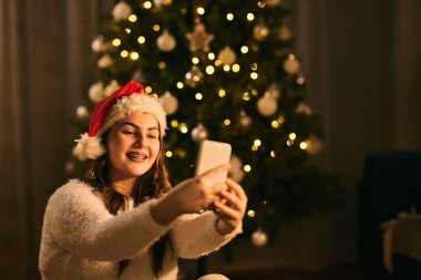 Noel Baba şapkalı genç bir kız güzel dekore edilmiş bir Noel ağacından selfie çekiyor.. 