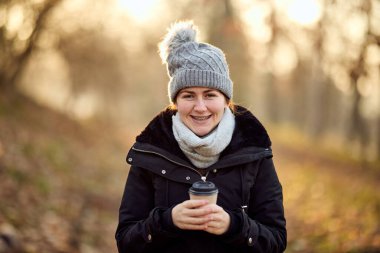 Sonbaharda kahvesini yudumlarken parkta yürüyen gülümseyen bir kadın.. 