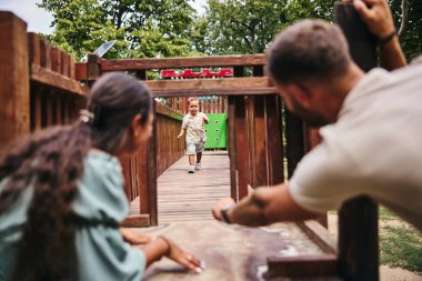 Küçük bir çocuk sevinçle gülen ebeveynlere doğru koşar ahşap bir oyun bahçesinde. 