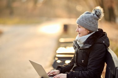 Şapkası ve atkısı olan mutlu bir kadın laptopunu doğada çalışmak için kullanıyor.. 