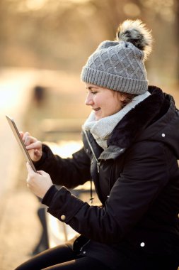 Gri şapkalı mutlu kadın güzel bir sonbahar gününde tabletini doğada kullanıyor.. 