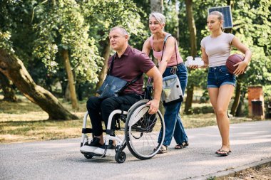Mutlu bir aile, parkta tekerlekli sandalyedeki bir adamla yürürken eğleniyor.. 
