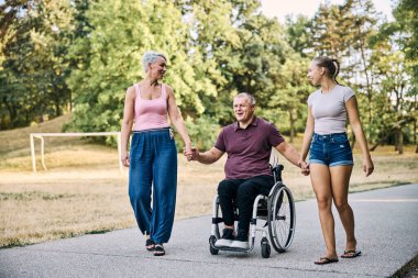 Tekerlekli sandalyedeki yaşlı bir adam ve parkta yürüyen iki kadından oluşan neşeli bir aile.,. 