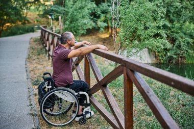 Tekerlekli sandalyedeki bir adam ahşap bir çitin yanında doğanın manzarasının tadını çıkarır..