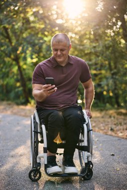 Tekerlekli sandalyedeki bir adam telefonunu açık havada kullanırken doğanın ve güneş ışığının tadını çıkarır..