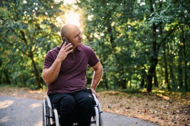 Tekerlekli sandalyedeki bir adam telefonunu açık havada kullanırken doğanın ve güneş ışığının tadını çıkarır..