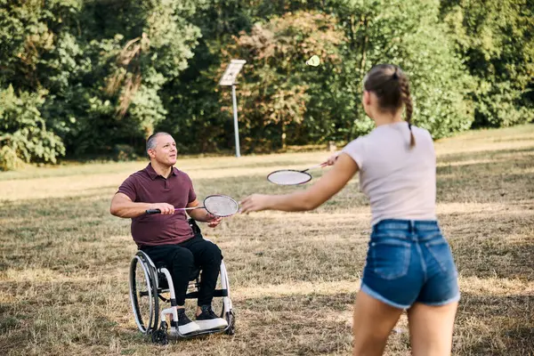Mutlu aile parkta tekerlekli sandalyedeki adamla badminton oynuyor.. 