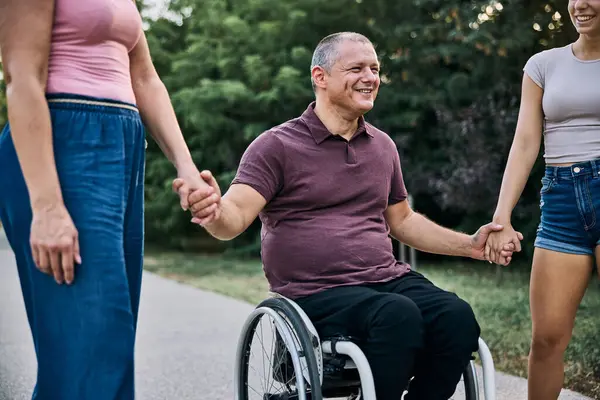 Tekerlekli sandalyedeki yaşlı bir adam ve parkta yürüyen iki kadından oluşan neşeli bir aile.,. 