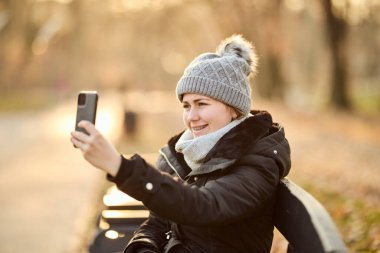 Şapkalı ve fularlı mutlu kadın bankta otururken fotoğraf çekiyor.. 