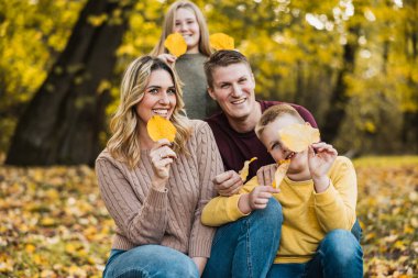 Dört kişilik bir aile, sonbahar parkında yapraklı poz veriyor ve onlarla oynuyor.. 