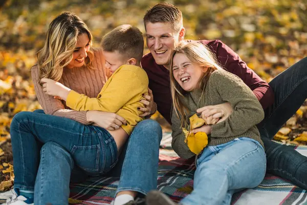 Dört kişilik neşeli bir aile güneşli bir sonbahar parkında battaniyenin üzerinde eğlenceli bir anı paylaşıyor.. 