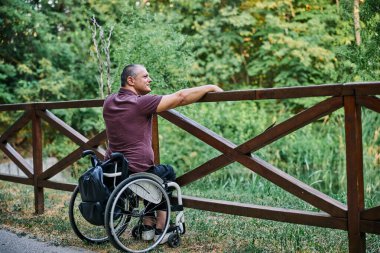 Tekerlekli sandalyedeki bir adam ahşap bir çitin yanında doğanın manzarasının tadını çıkarır.. 