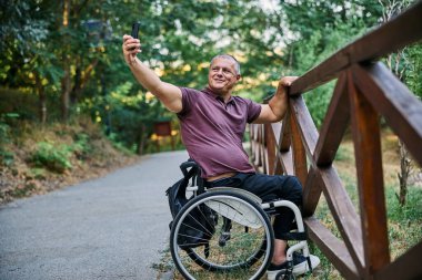 Tekerlekli sandalyedeki bir adam ahşap bir çitin yanında doğanın manzarasından zevk alır ve fotoğraf çeker.. 