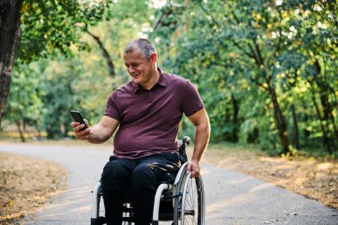Tekerlekli sandalyedeki bir adam telefonunu açık havada kullanırken doğanın ve güneş ışığının tadını çıkarır..