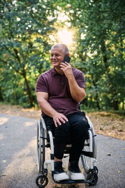Tekerlekli sandalyedeki bir adam telefonunu açık havada kullanırken doğanın ve güneş ışığının tadını çıkarır..