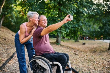Neşeli bir çift açık havada aşk ve arkadaşlığın altını çizerek selfie çeker. Adam tekerlekli sandalyededir., 