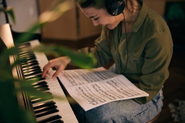 Bir kadın piyanonun başına oturur, notaları dikkatle okur, bir ev ortamında müzik odağı ve yaratıcılığını gösterir..