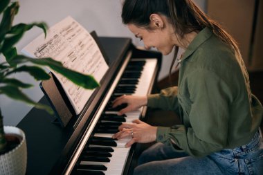 Genç bir kadın nota kağıdıyla piyano çalıyor, odak ve yaratıcılık anı yakalıyor..