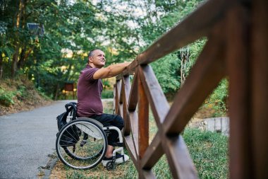 Tekerlekli sandalyedeki bir adam ahşap bir çitin yanında doğanın manzarasının tadını çıkarır..