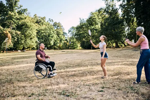 Mutlu aile parkta tekerlekli sandalyedeki adamla badminton oynuyor.. 