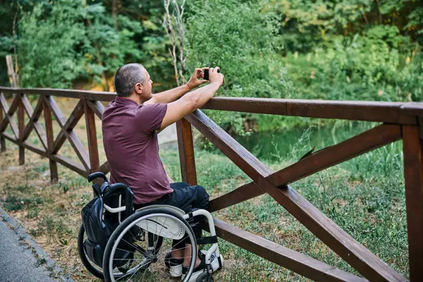 Tekerlekli sandalyedeki bir adam ahşap bir çitin yanında doğanın manzarasından zevk alır ve fotoğraf çeker.. 