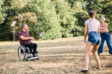 Mutlu aile parkta tekerlekli sandalyedeki adamla badminton oynuyor.. 