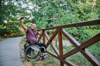 Tekerlekli sandalyedeki bir adam ahşap bir çitin yanında doğanın manzarasından zevk alır ve fotoğraf çeker.. 