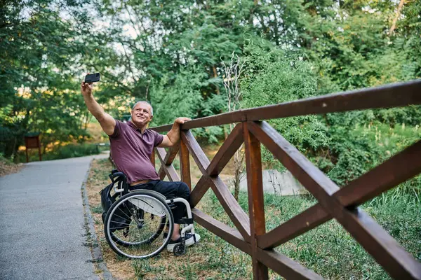 Tekerlekli sandalyedeki bir adam ahşap bir çitin yanında doğanın manzarasından zevk alır ve fotoğraf çeker.. 
