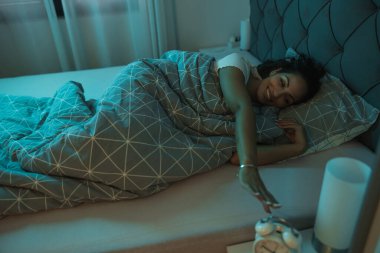 Bir kadın, sabahın erken saatlerinde battaniyenin altında yatağa uzanıp çalar saate uzanır.. 