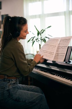 Bir kadın piyanonun başına oturur, notaları dikkatle okur, bir ev ortamında müzik odağı ve yaratıcılığını gösterir..