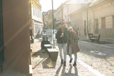 Neşeli bir çift manzaralı bir kaldırım taşı caddesinde birlikte yürüyor, sıcak güneş ışığı ve sonbahar titreşimlerinin tadını çıkarıyorlar..