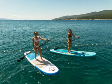 İki kadın güneşli bir gökyüzünün altındaki berrak mavi sularda kürek çekmekten hoşlanırlar.. 