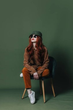 Stylish redhead woman in sunglasses and ginger shirt and pants sits in chair on green background