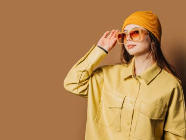 Stylish teenager girl in shirt and hat with sunglasses on brown background