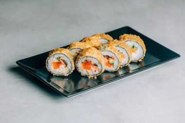 Kurutulmuş ekmek kırıntılarında sıcak suşi yuvarlanıyor. Gri arka planda siyah tabakta somon var.