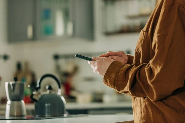 茶色のシャツの女性は台所で手にスマートフォンを保持し 表示するために探しています 背景にぼっけでキッチン付きサイドビュー ロイヤリティフリーのストック写真