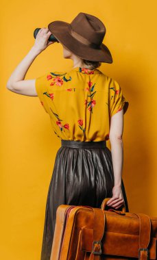 Şık, sarı saçlı, şapkalı, gömlekli, dürbünlü ve sarı arka planda bavulu olan kadın.