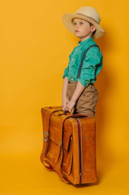 Şapkalı küçük çocuk, pantolon askısı ve sarı çantalı yeşil gömlek.