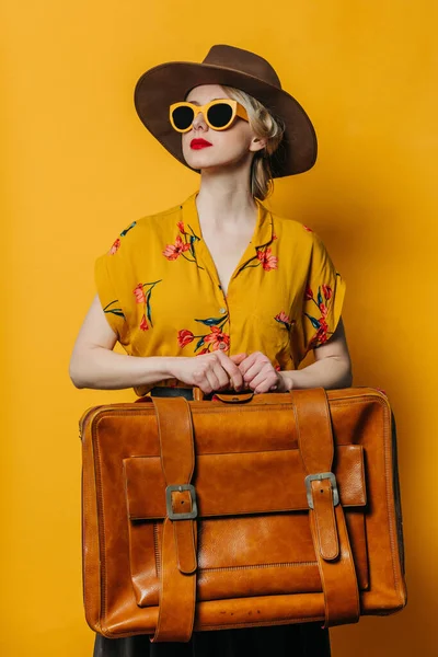 头戴帽子 戴着太阳镜 身穿短裙的金发女士 穿着古色古香的皮箱 背景是黄色的 — 图库照片
