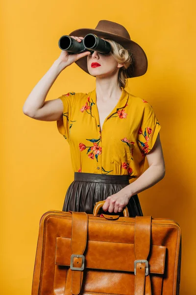 黄色の背景に双眼鏡とスーツケースと帽子とシャツのスタイリッシュなブロンドの髪の女性 — ストック写真