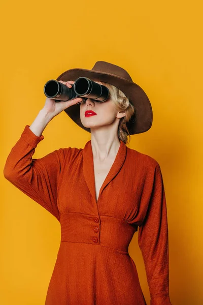黄色の背景に双眼鏡で帽子と茶色のドレスのスタイリッシュなブロンドの髪の女性 — ストック写真