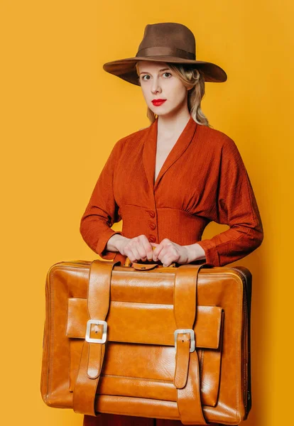 ブロンド髪女性でヴィンテージ帽子と茶色のドレスとともにスーツケース上の黄色の背景 — ストック写真