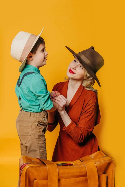 Stylish mother and son in vintage clothes with suitcase on yellow background