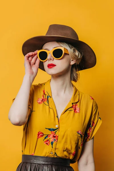 黄色の背景にスカートと帽子 サングラスやシャツのスタイリッシュなブロンドの髪の女性 ストック画像