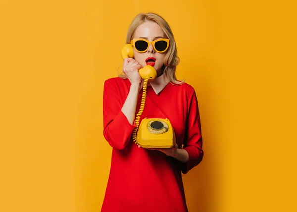 黄色の背景にダイヤル電話で赤いドレスでスタイリッシュな驚きの女性 — ストック写真