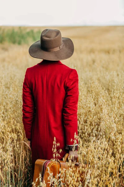 田舎のオートフィールドのスーツケースとヴィンテージ帽子の赤いコートのスタイリッシュな女性 — ストック写真