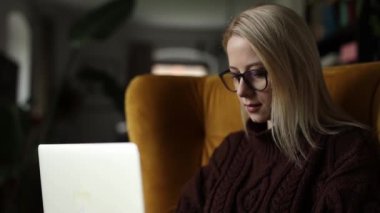 Gözlüklü ve laptoplu beyaz kadın evdeki koltukta oturuyor.